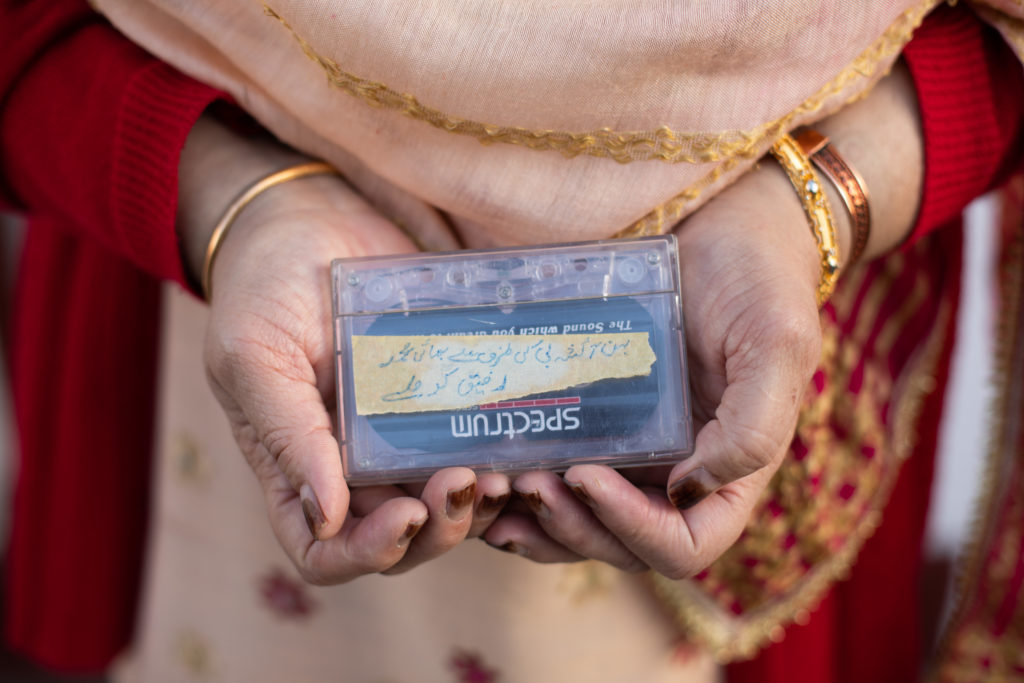 The hands of a woman holding a cassette tape, wearing gold bracelets and cream and red clothes.