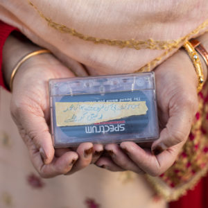 The hands of a woman holding a cassette tape, wearing gold bracelets and cream and red clothes.