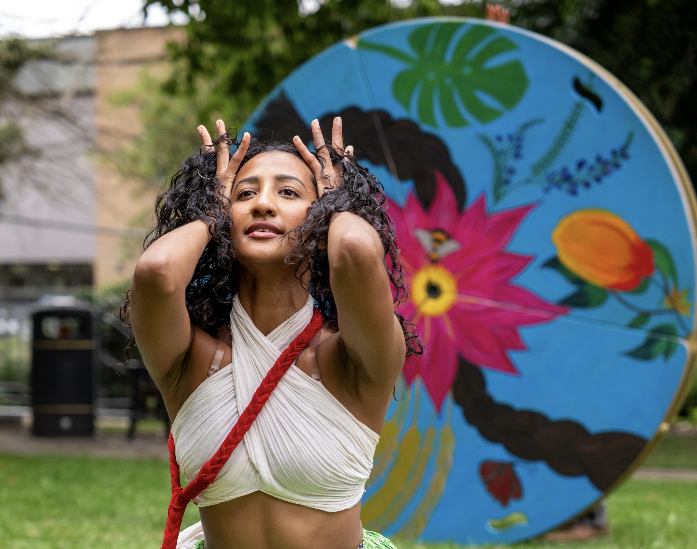 In a natural setting, a South Asian woman in white blouse with a plaited red sash across her body, has her hands and fingers moving upwards through her open curly black hair. Her eyes are knowing and hopeful. Behind her is a brightly painted coloured wheel including a plait of hair, a pink lotus flower, a mango, a green leaf, a butterfly, an indigo plant, wheat sheaths and a bee.