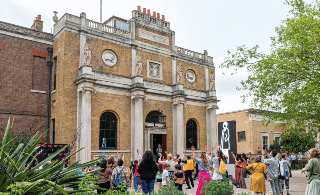 Visitors of all ages gather outside the front of Pitzhanger Manor, raising their hands as they take part in a lively outdoor performance during a community event, with the historic façade and contemporary gallery entrance in the background.